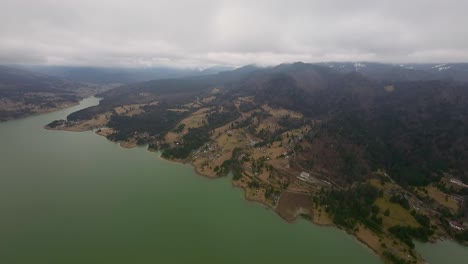 Aerial-view-of-wonderful-Colibita-lake-along-forested-hills-with-scattered-rural-homes-on-a-cloudy-day-in-Transilvania,-Romania