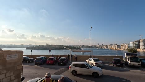 La-Vista-Panorámica-Del-Puerto-De-La-Valeta-En-Malta,-Capturada-Desde-La-Bulliciosa-Zona-De-Estacionamiento-Con-Peatones-Y-Viajeros,-Muestra-La-Vibrante-Vida-Marítima-Y-La-Atmósfera-Urbana.