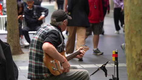 Talented-musician-busks-on-Bourke-Street-Mall,-playing-the-guitar-and-singing-in-the-heart-of-bustling-Melbourne-city,-with-pedestrian-passing-by-in-the-scene