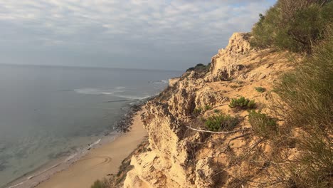 La-Escarpada-Costa-De-España-Muestra-Una-Playa-De-Arena-Junto-A-Acantilados-Sedimentados,-Todos-Iluminados-Por-El-Brillante-Sol-Diurno,-Capturando-La-Esencia-De-La-Belleza-Costera-Y-Las-Maravillas-Naturales.