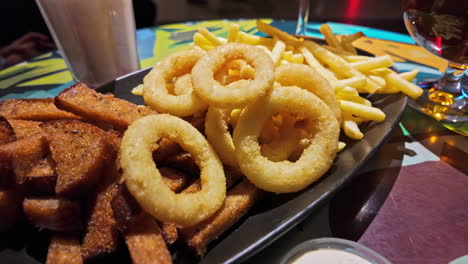 Snack-plate-with-onion-rings,-french-fries-and-bread-in-a-restaurant-inside-and-sauces-and-drinks-around-close-up