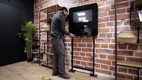 Man-assembling-a-modern-black-screen-stand-in-a-stylish-room-with-brick-walls-and-green-plants