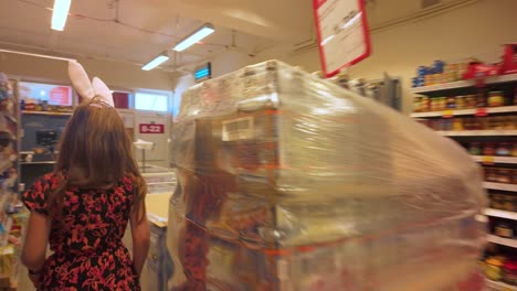 Girl-wearing-red-flower-dress-and-rabbit-ears-walks-through-local-supermarket,-seen-from-the-rear-view