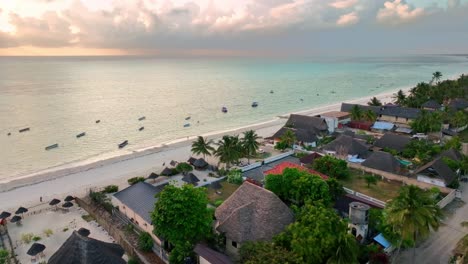 Drone-Se-Eleva-Desde-El-Océano-Para-Capturar-Paje,-Zanzíbar:-La-Puesta-De-Sol-Dorada-Ilumina-La-Playa-Y-El-Paisaje-Urbano