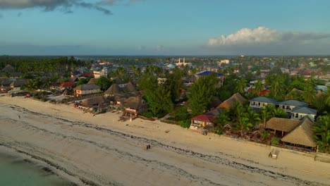 "Watch-as-the-drone-captures-Paje,-Zanzibar-at-sunset:-aerial-view-of-beach,-cityscape,-and-golden-hour-glow