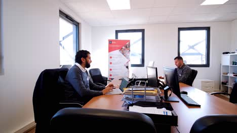 Dos-Hombres-Trabajando-En-Escritorios-En-Una-Oficina-Moderna-Y-Luminosa-Con-Grandes-Ventanales