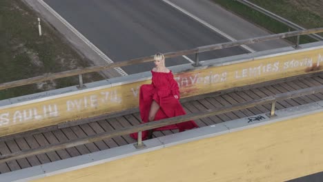 Eine-Drohne-Fliegt-Von-Einem-Model-Mit-Rotem-Kleid-Weg,-Das-Auf-Einer-Brücke-Posiert