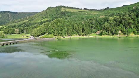 Lush-green-hills-reflected-in-a-calm-lake-surrounded-by-trees-and-a-bridge