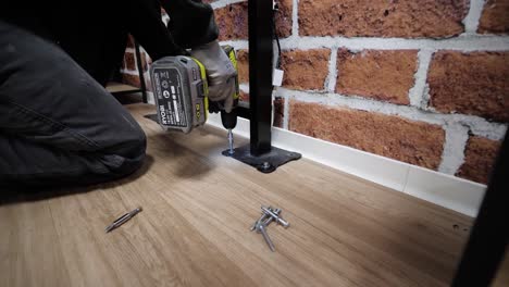 Close-up-of-a-person-using-a-power-drill-to-secure-a-stand-to-a-wooden-floor-beside-a-brick-wall