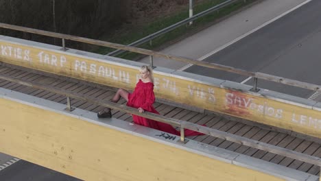 Eine-Drohnenaufnahme-Eines-Models-Im-Roten-Kleid,-Das-Sein-Bein-Auf-Eine-Brücke-Stellt