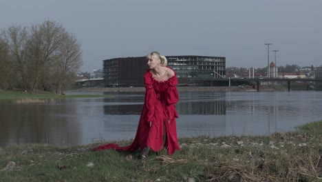 Una-Impresionante-Toma-En-Cámara-Lenta-De-Una-Mujer-Con-Un-Vestido-Rojo-Brillante-Cerca-Del-Río-Posando