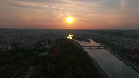 Aerial-drone-flyover-Prague-at-sunset-showcases-the-serene-Vltava-River,-historical-buildings,-the-golden-sun-casts-a-warm-glow,-reflecting-off-the-water-and-creating-a-picturesque-urban-landscape