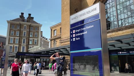 Turistas-Que-Entran-En-La-Estación-Kings-Cross-En-Londres-Con-Un-Cartel-Azul-De-Kings-Cross-Square-En-Primer-Plano-En-Un-Día-Soleado