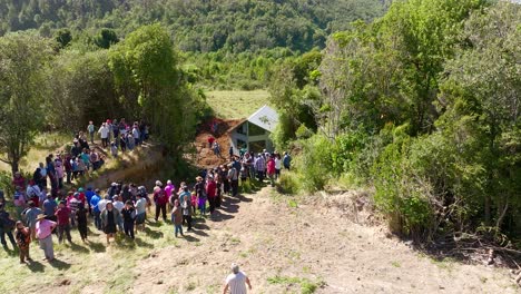 A-large-group-of-people-gathered-to-witness-the-relocation-of-a-house-aided-by-heavy-machinery-in-a-rural,-lush-green-landscape-in-Chiloe