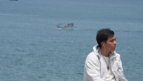 young-model-asiatic-guy-listens-to-music-with-boat-sealing-the-sea-ocean-in-background-,-portrait-lifestyle-in-slow-motion