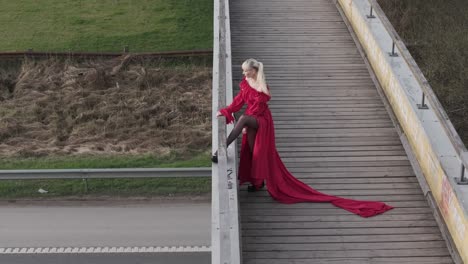 Eine-Drohnenaufnahme-Mit-Einem-Model-In-Einem-Leuchtend-Roten-Kleid,-Das-Mit-Den-Beinen-Auf-Einer-Brücke-Posiert