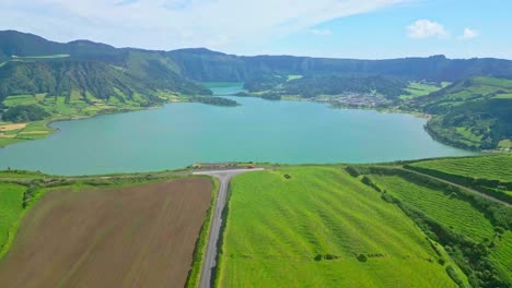 A-serene-lake-surrounded-by-lush-green-hills-and-a-distant-village,-aerial-view