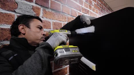 Close-up-of-a-man-using-a-power-drill-to-work-on-a-black-surface-next-to-a-brick-wall