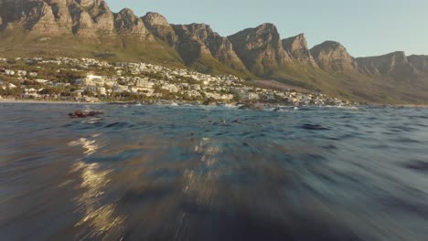 El-Dron-Vuela-Rápido-Sobre-El-Mar-Ondulado-En-La-Playa-De-Camps-Bay-En-Ciudad-Del-Cabo,-Sudáfrica---Muchas-Casas-En-La-Cima-De-Una-Colina---Vista-De-Las-Rocas-De-La-Montaña-De-La-Mesa-En-El-Mar---El-Dron-Vuela-A-Través-De-Un-Enjambre-De-Aves