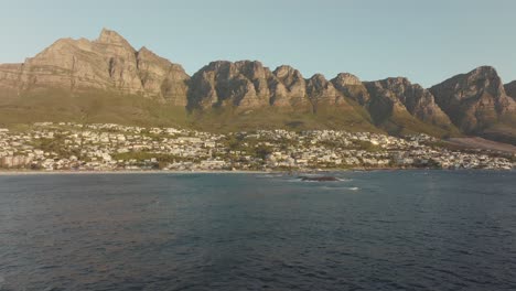 Drone-flies-high-backwards-over-the-undulating-sea-at-Camps-Bay-beach-in-Cape-Town-South-Africa---many-houses-on-a-hilltop---view-of-Table-Mountain-rocks-in-the-sea