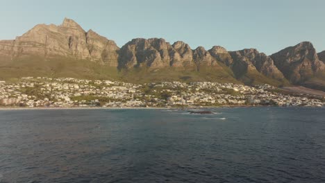 Drone-Vuela-Alto-Hacia-Atrás-Sobre-El-Mar-Ondulado-En-La-Playa-De-Camps-Bay-En-Ciudad-Del-Cabo,-Sudáfrica---Muchas-Casas-En-La-Cima-De-Una-Colina---Vista-De-Las-Rocas-De-La-Montaña-De-La-Mesa-En-El-Mar