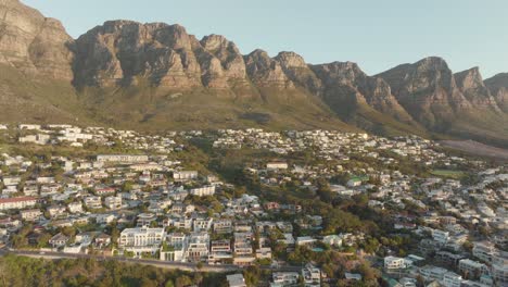 Drone-flying-high-backwards-on-top-of-camps-bay-in-cape-town-south-africa---many-houses-on-a-slope---view-of-table-mountain-and-the-wide-sea-at-sunset