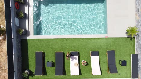 Drone-flies-over-an-outdoor-pool-in-bird's-eye-view---Woman-in-swimming-costume-gets-out-of-the-pool-and-lies-on-sun-lounger---Villa-in-Greece,-Crete-with-palm-trees