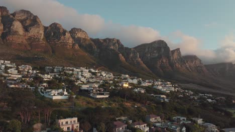 Drones-Volando-Lentamente-De-Lado-Sobre-La-Bahía-De-Camps-En-Ciudad-Del-Cabo,-Sudáfrica---Vista-De-La-Montaña-De-La-Mesa-Al-Atardecer-Con-Nubes-Claras