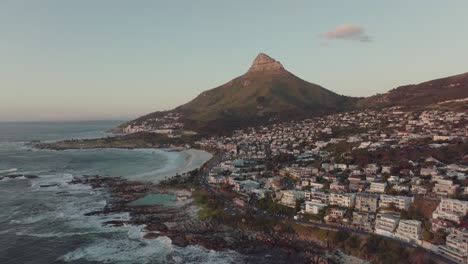 El-Dron-Vuela-Alto-Y-De-Lado-Sobre-La-Playa-De-Camps-Bay-En-Ciudad-Del-Cabo,-Sudáfrica:-La-Montaña-De-Cabeza-De-León-Iluminada-Por-El-Atardecer-Se-Eleva-Al-Fondo