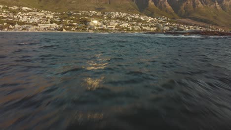 Drone-flies-fast-over-the-wavy-sea-at-Camps-Bay-beach-in-Cape-Town-South-Africa---many-houses-on-a-hilltop---view-of-table-mountain-rocks-in-the-sea---birds-fly-by