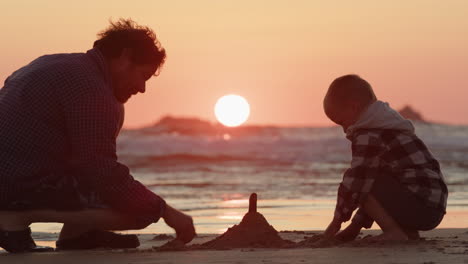 Single-dad-and-son-builds-sandcastle-on-beach-with-vibrant-sunset-between-them