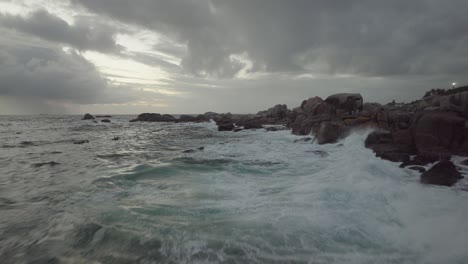 Drohne-Fliegt-Rückwärts-Knapp-über-Dem-Meer-Am-Camps-Bay-Beach-In-Kapstadt,-Südafrika-–-Es-Ist-Ein-Bewölkter-Tag-Und-Die-Wellen-Schlagen-Gegen-Die-Felsen