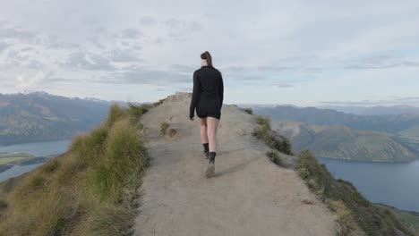 Young-caucasian-woman-with-ponytail-and-in-shorts-walking-to-a-viewpoint-at-Roy's-Peak,-looking-at-Lake-Wanaka-and-mountains-in-Wanaka,-New-Zealand