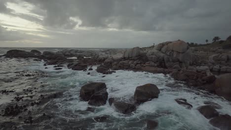 Drone-flying-in-reverse-just-above-the-sea-at-Camps-Bay-beach-in-Cape-Town-South-Africa---It's-overcast-and-the-waves-are-crashing-against-the-rocks