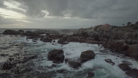 Drone-Volando-En-Reversa-Justo-Sobre-El-Mar-En-La-Playa-De-Camps-Bay-En-Ciudad-Del-Cabo,-Sudáfrica:-Está-Nublado-Y-Las-Olas-Rompen-Contra-Las-Rocas