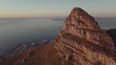 Drone-flies-high-at-the-Lion's-Head-mountain-peak---The-rocks-are-illuminated-by-the-sunset---In-the-valley-you-can-see-Camps-Bay-beach-in-Cape-Town-South-Africa---view-of-the-open-sea