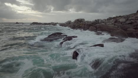 Drone-Volando-Justo-Sobre-El-Mar-En-La-Playa-De-Camps-Bay-En-Ciudad-Del-Cabo,-Sudáfrica:-Es-Un-Día-Nublado-Y-Las-Olas-Rompen-Contra-Las-Rocas