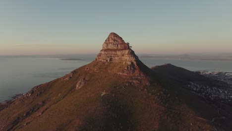 Drone-flies-slowly-towards-the-Lion's-Head-mountain-peak---They-are-illuminated-by-the-sunset---In-the-valley-you-can-see-Cape-Town-South-Africa---view-of-the-open-sea