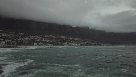 Drone-flies-just-above-rocks-at-Camps-Bay-beach-in-Cape-Town-South-Africa---The-sea-has-high-waves-and-in-the-background-you-can-see-Table-Mountain-covered-in-clouds