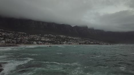 El-Dron-Vuela-Justo-Encima-De-Las-Rocas-En-La-Playa-De-Camps-Bay-En-Ciudad-Del-Cabo,-Sudáfrica:-El-Mar-Tiene-Olas-Altas-Y-Al-Fondo-Se-Puede-Ver-La-Montaña-De-La-Mesa-Cubierta-De-Nubes.