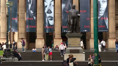 The-State-Library-Victoria-with-Corinthian-columns,-featuring-the-statue-of-Sir-Redmond-Barry-located-on-the-forecourt,-people-hanging-out-on-the-lawn-and-pedestrians-walking-on-the-sidewalks