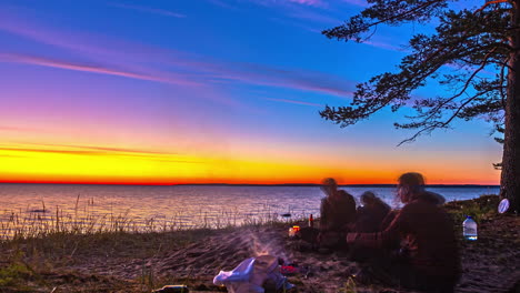 People-wild-camping-by-the-sea-with-campfire,-early-morning-sunrise-time-lapse