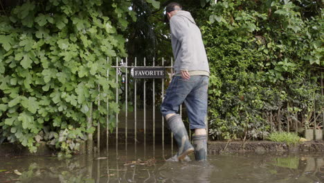 Un-Hombre-Con-Botas-De-Lluvia-Ingresa-A-La-Propiedad-Desde-Una-Calle-Inundada-A-Través-De-La-Puerta-Del-Jardín