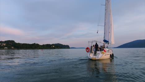 Vista-Aérea-De-Un-Velero-Navegando-Por-Aguas-Tranquilas-Durante-La-Puesta-De-Sol,-Con-Terreno-Montañoso-Al-Fondo-Y-Suaves-Ondulaciones-En-La-Superficie-Del-Lago