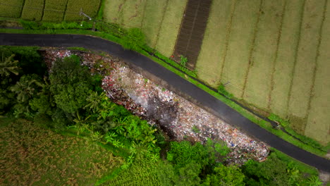 Aerial-view-of-smoldering-garbage-pile-next-to-country-road-and-green-farm-land