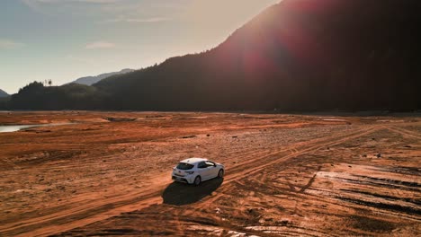 white-Toyota-Corolla-navigates-the-rugged,-bumpy-terrain-of-the-mud-flats-in-the-dried-up-bed-of-Stave-Lake-in-Mission-BC-Canada-The-car-kicks-up-clouds-of-brown-sand-rock-dirt-and-dust-as-it-moves