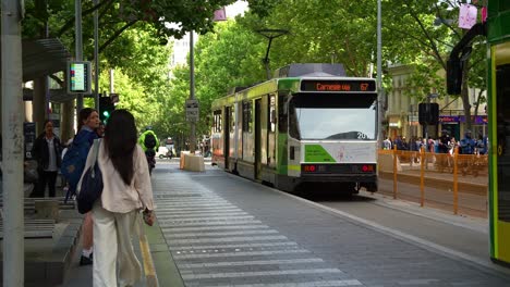 Cyclists-and-scooter-ride-through-the-lane-and-trams-glides-along-tree-lined-Swanston-street-with-commuters-waiting-at-the-stop,-a-vibrant-scene-of-Melbourne's-bustling-Central-Business-District