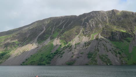 Cloud-shadows-moving-up-steep-scree-sided-mountain-next-to-lake-with-small-boat-at-Wastwater