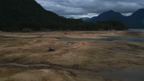 Drone-flies-fast-in-reverse-tracking-black-pickup-truck-parked-on-muddy-flats-starts-medium-wide-close-extends-to-high-angle-far-back-revealing-dark-green-mountains-trees-blue-clouds-vehicle-tracks