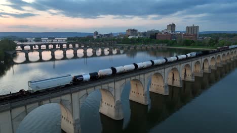 Aufsteigende-Drohnenaufnahme-Eines-Güterzuges,-Der-Güter-Auf-Einer-Brücke-über-Den-Susquehanna-River-Transportiert,-Während-Des-Goldenen-Sonnenuntergangs